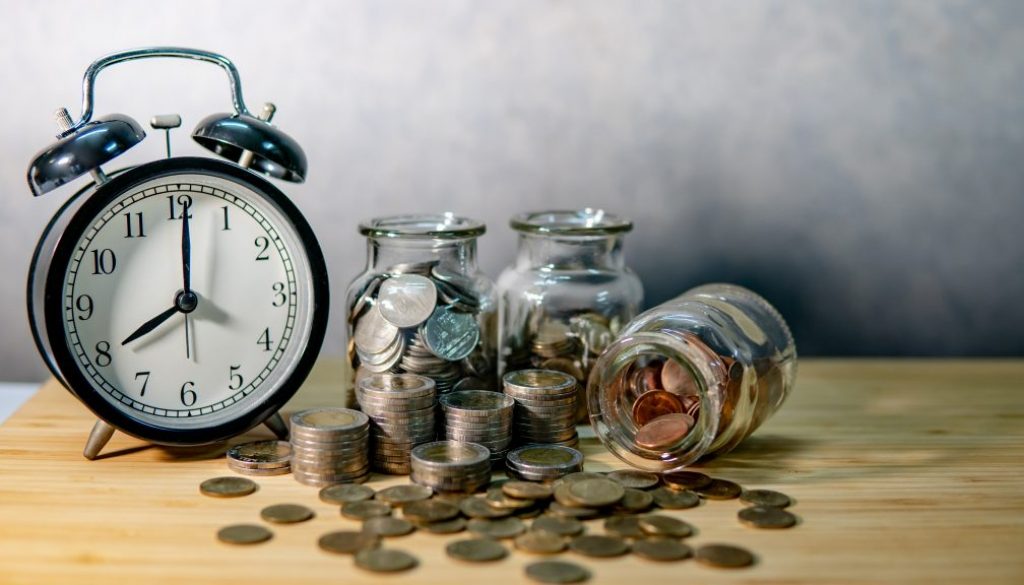 Clock with coins in currency glass jars and spilling on wooden table. Saving money for future retirement. Financial business growth. Time investment concept