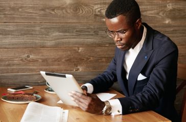 Serious young African American businessman holding digital tablet, looking at the screen with concentrated expression while filling in some papers, sitting at a cafe, having coffee during lunch break