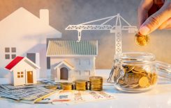 Real estate or property development. Construction business investment. Home mortgage loan rate. Hand putting coin in currency glass jar with Coin stack, banknotes, house and crane models on the table.