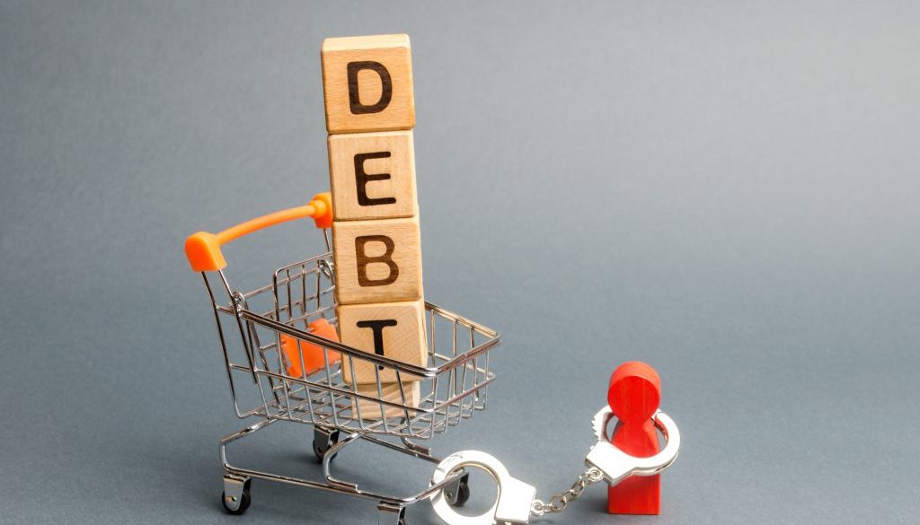 Wooden blocks with the word Debt in a supermarket trolley and a human figurine bound by handcuffs. Mandatory payment of debt. Alimony payment. Pay of a mortgage loan. Debt load / obligation. IOU